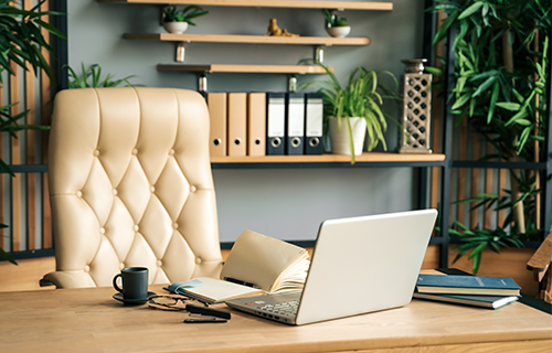 desk setting with chair, coffee cup, open laptop, notebooks, and green plants in the background