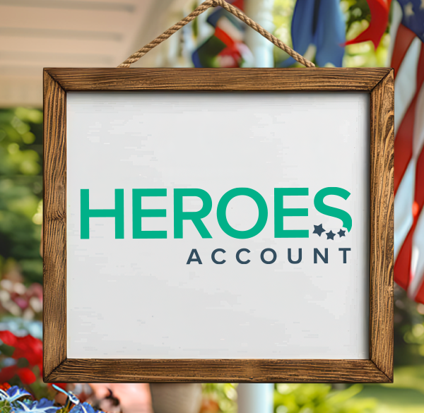 Wooden frame with Heroes Account written in green and navy on white background with the frame hanging from porch with American flag in the back with red, white, and blue flowers on the lefthand side blurred